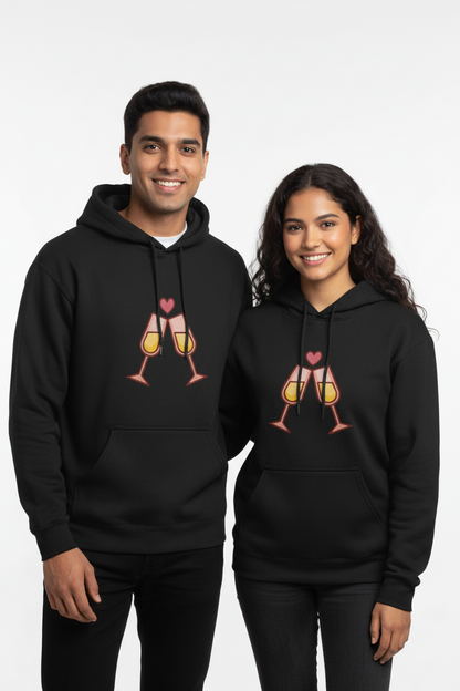 Smiling young man and woman wearing matching black hoodies with clinking champagne glasses and heart graphic